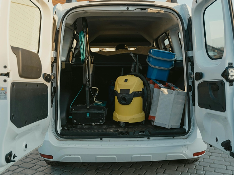 Vue de l’arrière d’un véhicule utilitaire blanc avec les portes ouvertes, contenant du matériel de nettoyage professionnel, dont un aspirateur industriel jaune, des seaux bleus empilés, une serpillière avec manche, des caisses métalliques et divers équipements.