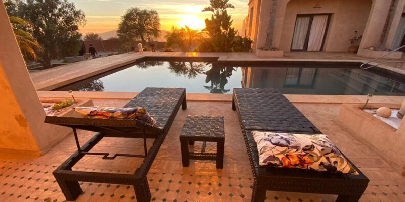 Terrasse avec transats face à une piscine privée, au coucher du soleil, dans une villa marocaine entourée de palmiers et de verdure.