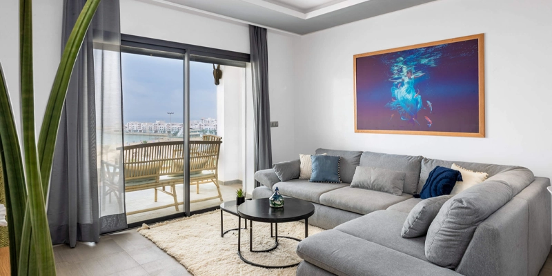 Salon moderne et lumineux dans un appartement à Agadir, avec canapé d’angle gris, grande baie vitrée donnant sur une terrasse avec vue sur la ville.