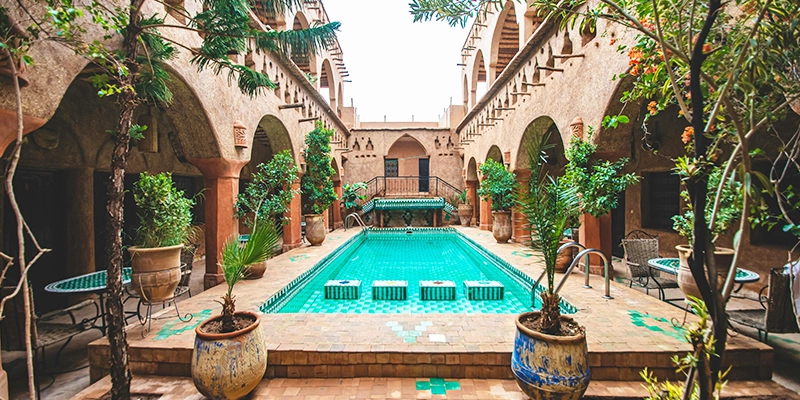 Patio traditionnel marocain avec piscine centrale en carreaux verts, entouré d’arches, de plantes et de salons extérieurs.